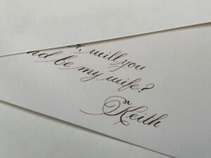 Close-up of handwritten wedding invitation text reading, “Will you be my wife?” with an elegant signature below. Calligrapher, Jagdeep Sahans